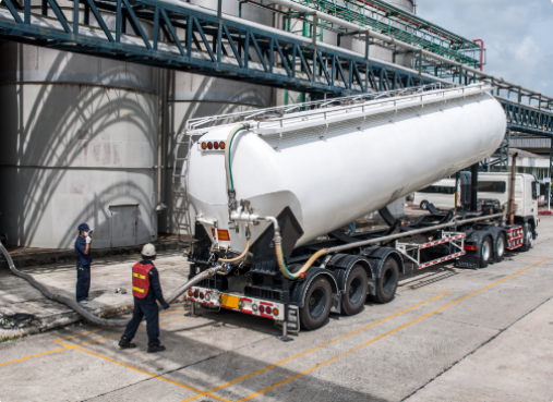 Two workers in safety gear monitor the unloading of a large white tanker truck at an industrial facility